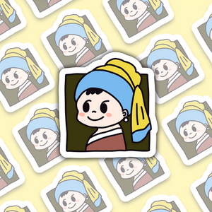 Hand holding a small cartoon sticker inspired by "Girl with a Pearl Earring," featuring a cute character with a blue headscarf and yellow details, against a blurred indoor background.