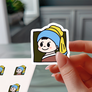 Hand holding a small cartoon sticker inspired by "Girl with a Pearl Earring," featuring a cute character with a blue headscarf and yellow details, against a blurred indoor background.