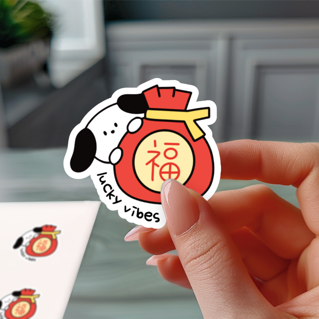 A cute vinyl sticker featuring a black and white cartoon dog hugging a red lucky money pouch with the Chinese character "福" (fu, meaning fortune or blessing). The words “lucky vibes” are written underneath in playful, handwritten text
